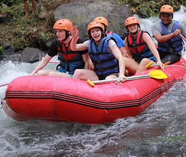 Excited tourists white water rafting in a red boat with life jackets and helmets on a tropical river.