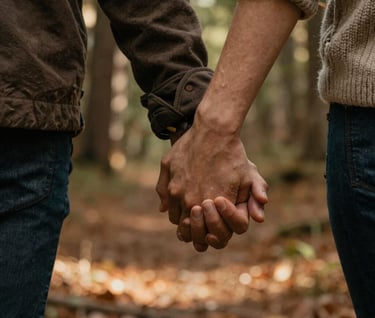 A close-up, cinematic detail shot of two people holding hands during a walk in a North American / US forest. Warm sunbeams filter through the trees. The light highlights textures of soft wool and skin. Earthy Muted Brown and Terracotta accents in the background foliage.