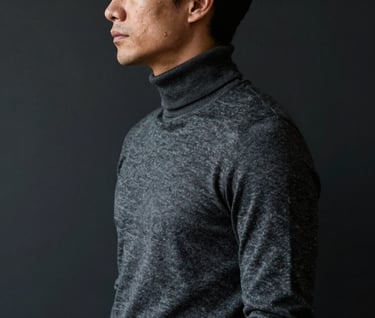 A profile view of a man in a dark slate grey turtleneck, standing against a deep charcoal black wall. Cinematic lighting creating a sharp silhouette.