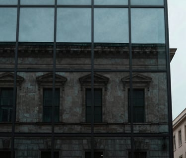 A minimalist portrait of a historic building facade reflected in a modern glass window. The contrast between old and new is captured in shades of Muted Teal and Dark Slate under a soft, overcast sky.