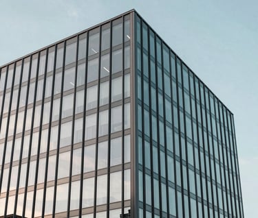 An architectural photograph of a minimalist International / Western glass building reflecting a soft sky blue sky. The image emphasizes clean lines and modern structural precision.