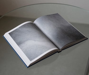 An independent photography book lying open on a minimalist glass table. The pages show abstract soft gray patterns. Muted steel blue accents. Global / Contemporary Art aesthetic.