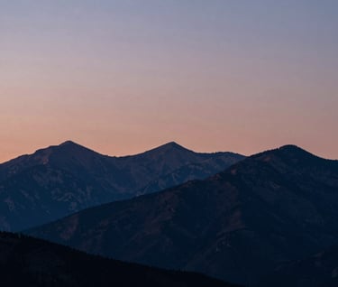 An artistic landscape of a North American / US mountain range at twilight. The sky is a gradient of dusty rose and muted blue, with the silhouettes of peaks in deep navy.