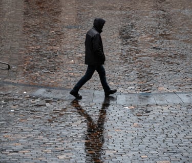 A street photography scene in Utrecht. A single figure walking across a cobblestone square in the rain. Reflective charcoal pavement, soft light, minimalist composition capturing a quiet, real moment in a Northern European city.