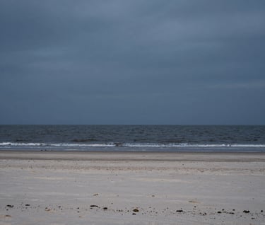A cinematic photograph of a deserted beach on the North Sea at dusk. The sand is a pale grey-beige, meeting the charcoal tide. Wide, minimalist composition with a vast, heavy sky. Premium storytelling photography.