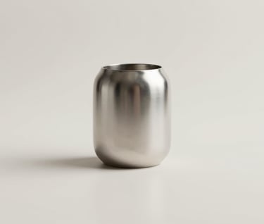 A minimalist arrangement of a single metallic object reflecting soft light. The background is a clean, uniform #F8F5F2. The aesthetic is 'quiet luxury', focusing on material quality and simple forms.