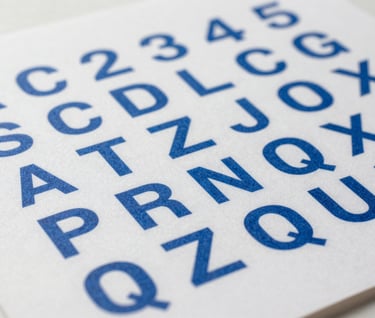 Macro photography of a high-quality art print showing a grid of geometric alphabet characters in cerulean blue. The shot is taken in a brightly lit North American studio with soft shadows and sharp focus on the paper texture.