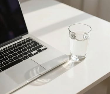 A clean and professional workspace with a sleek laptop and a glass of water on a white desk. Sunlight creating soft natural shadows. International / Professional. Colors: white, off-white, and light grey.