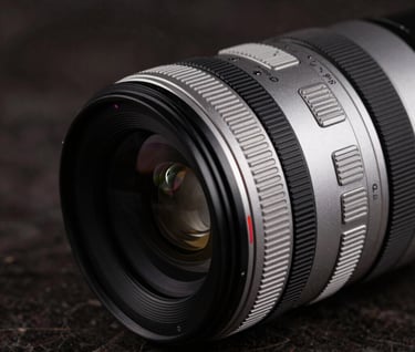 A detailed close-up of a modern camera lens sitting on a dark marble surface. The lighting is low-key and dramatic, picking up the silver and black metallic textures. International / Professional aesthetic.