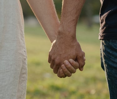 A close-up, shallow depth-of-field photograph of two people holding hands while walking in a North American park. The light is warm late-afternoon golden. Focus on the authentic connection and skin texture.