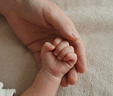 A detailed close-up shot of a parent's hand gently holding a newborn's tiny hand, set against a soft, light beige knit blanket. Captured in North American / US (West Michigan) with warm, natural morning light.