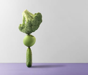 a head of broccoli balanced on a green apple and pickle balanced on a green cucumber