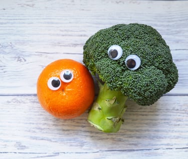 a broccoli and an orange with plastic eyes are cuddled up together on a table