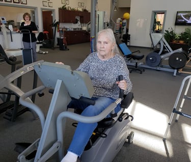 a woman in a gym room with a man on a stationary bike