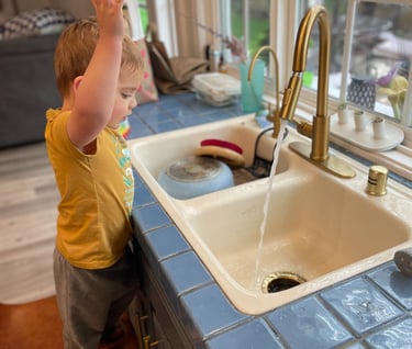 little boy standing on chair at kitchen sink with hands in the air