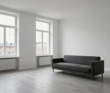 A wide-angle interior photograph of a minimalist residential space in Stockholm. The room features white walls, light gray flooring, and a single piece of dark gray furniture. Large windows invite soft, diffused Northern European daylight, creating a serene and professional atmosphere.