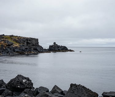 A wide-angle landscape photograph of a rugged Northern European coast, featuring dark charcoal volcanic rocks meeting a calm, light gray sea under a misty sky. The composition is minimalist and serene, emphasizing vast, quiet space.