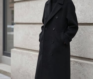 A professional editorial fashion photograph of a person wearing a tailored black wool coat, standing against a minimalist light gray stone wall in a Northern European city. The lighting is soft and overcast, highlighting the clean silhouette and sophisticated mood.