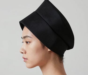 Close-up of a model's profile wearing a bold, geometric headpiece in black #0F0F0F. Sharp focus on the craftsmanship, against a light gray #D0D0D0 background. Artistic and refined.