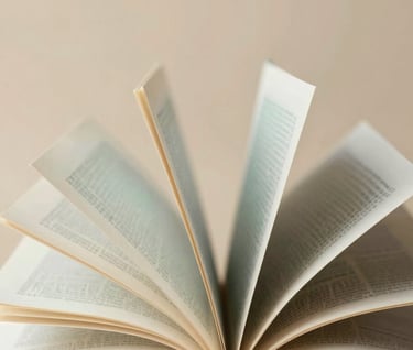 A creative arrangement of open books where the pages are fanned out artistically, set against a warm sandy background #D4B281. The book covers feature muted teal #7A9E9F and charcoal #3D3B3C tones.