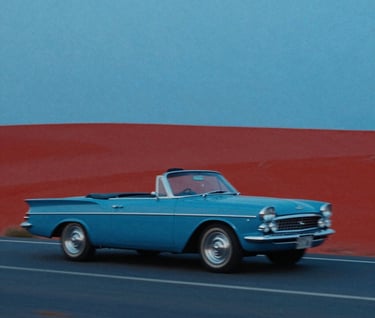 A cinematic wide shot of a 1960s convertible car driving through a landscape of flat #0050B3 blue and #D4202B red shapes. The composition is extremely asymmetric, following Godard's visual style. Heavy film grain and a sense of motion blur in a graphic, non-realistic way.