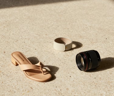 A high-fashion still life photograph showcasing handcrafted personal accessories on a solid travertine stone surface. Warm side lighting, editorial magazine layout with generous negative space, neutral sand and cream color tones.