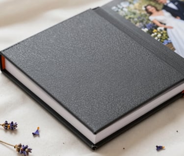 Close-up of a beautifully designed wedding photo album with a textured charcoal cover, resting on a soft cream fabric with a few lavender petals scattered around.