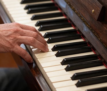 A detailed close-up of a hand resting on the keys of an antique upright piano in a North American / US music hall. The image has a timeless, elegant feel. The keys are a soft pale cream and the wood is a rich dark charcoal brown.