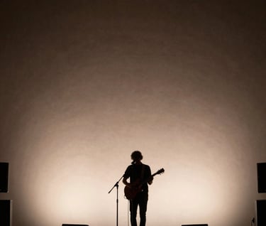 A wide-angle shot of a musician performing on a small, intimate stage in a North American / US lounge. The atmosphere is filled with a soft hazy light. The background is a dark charcoal brown, with the performer silhouetted against a pale cream light.