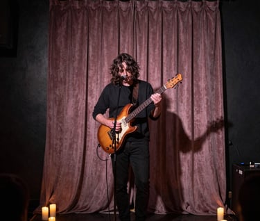 A musician performing on a small stage in a candlelit North American / US club. The lighting creates an intimate glow against a dark charcoal background. The mood is artistic and soulful, with muted mauve hues in the stage curtains.