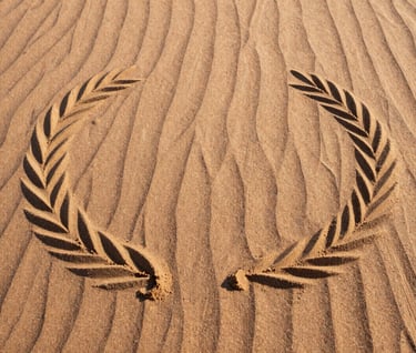 A top-down view of a sand art pattern resembling ripples on water, created on a warm desert surface. The composition is minimal and elegant, featuring a palette of soft sand and deep charcoal shadows cast by the afternoon sun.