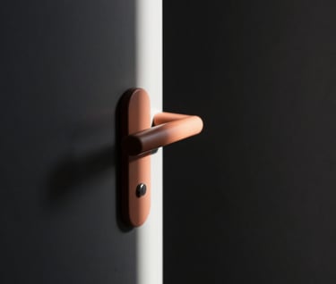 An abstract architectural study focusing on the play of light. A thin strip of light cutting through a dark charcoal (#333333) hallway, illuminating a terracotta (#BC6C47) door handle.