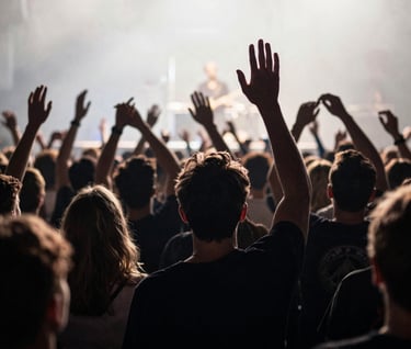 An atmospheric shot from the back of a concert crowd, showing blurred silhouettes of fans with hands raised toward a stage lit in cool off-white and charcoal tones. Capturing the collective emotion of live music in a Western European / Dutch setting.