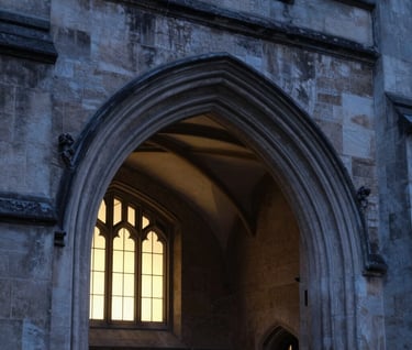 An architectural detail of a Gothic stone archway at a prestigious university during the blue hour. The textures of the charcoal-colored stone are sharp and detailed, with cream light spilling from a window in the background.