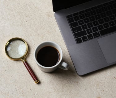 A top-down view of a minimalist workspace. A dark charcoal laptop sits next to a small ceramic cup of espresso and a single, antique gold-rimmed magnifying glass. The surface is a textured cream stone.