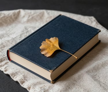 A still life photograph of a heavy hardcover book resting on a soft cream linen cloth. A single dried ginkgo leaf in antique gold serves as a bookmark. The background is a deep charcoal, creating a moody, literary feel.