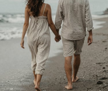 A candid, grain-textured photo of a couple walking barefoot along a quiet Bali beach, their backs to the camera, soft wind blowing through linen clothing, muted and nostalgic #D4C7BB color palette.