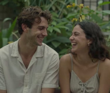 A candid moment of a couple sharing a quiet laugh in a lush green garden. The style is cinematic and story-driven, with subtle earth tones like #8F6E5F in the shadows.