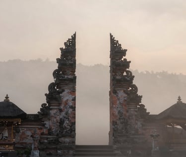 Wide shot of a misty Balinese temple at dawn. Ethereal, quiet, and peaceful atmosphere. Soft #D4C7BB and #F8F4F0 hues in the morning sky.