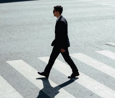 A candid street photograph of a person walking across a zebra crossing in a city. High contrast lighting with long shadows on light gray-blue pavement. Minimalist style, International / Western urban environment.