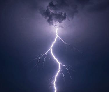 Powerful lightning photography showing a single, electric white bolt striking across a dark navy blue sky over a distant, flat horizon. The composition is elegant and stark, emphasizing the raw energy of the storm.