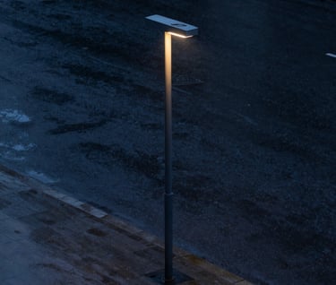 A minimalistic street life photograph during a rainy evening. A single modern street lamp illuminates wet, dark navy pavement. The composition is clean and empty, emphasizing solitude and elegance. International / Western urban environment.