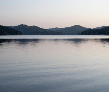 Minimalistic landscape photography of a calm lake at dawn. The water is a smooth, silvery blue-grey, reflecting the silhouette of distant hills under a soft, overcast sky. Pure simplicity and elegance.