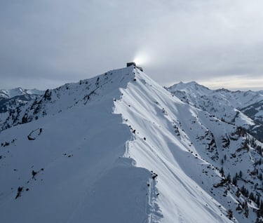 An expansive landscape photograph of a snowy mountain ridge in North America / US under a pale smoke winter sky. In the distance, a small, glowing charcoal black structure is perched on a cliff, casting a soft silver light into the mist.