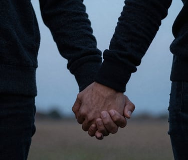 A close-up, storytelling photograph of two hands intertwined, belonging to a North American / US couple. They are standing in a field at dusk. The lighting is dim and cinematic, featuring muted light blue tones of the twilight sky and the soft charcoal black of their sweaters.