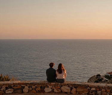 A wide, cinematic landscape photograph of a couple sitting on a rustic stone ledge overlooking the sea in Spain / Southern Europe. Soft evening light casting gentle orange, sand, and terracotta tones over the entire landscape. Peaceful, nostalgic, authentic photography.