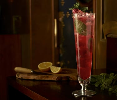 Red cocktail in a tall glass on a wooden table with mint and cut lemons
