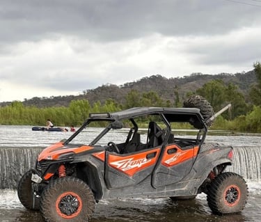 Orange CFMOTO ZFORCE side-by-side UTV parked in a shallow river by a waterfall under cloudy skies.