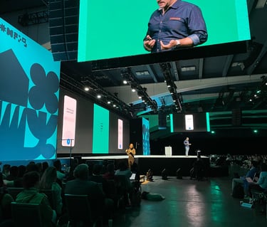large conference audience facing an ASL interpreter standing under a large screen with the speaker