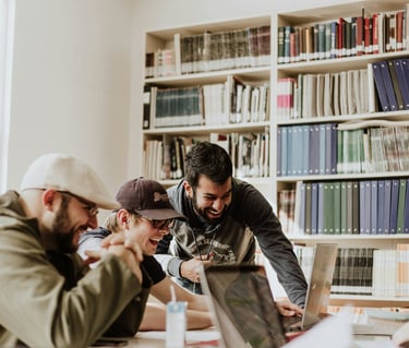 A group of diverse university students laughing and studying together with laptops in a bright library.
